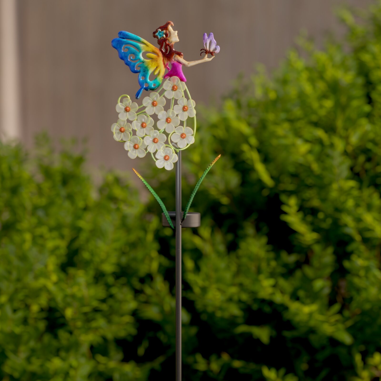 Kleurrijke decoratieve tuinprikkers in de vorm van feeën, met vlindervleugels en witte bloemen op een metalen staafje, zijaanzicht in de tuin tegen een groene achtergrond