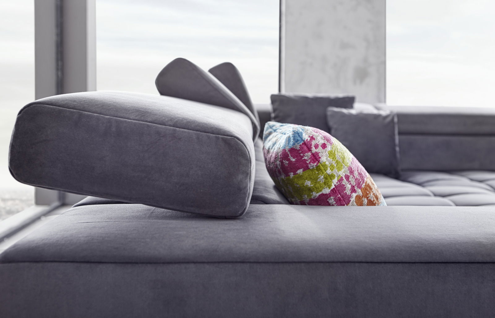 Nahaufnahme eines modernen Ecksofas in Grau mit verstellbarer Kopfstütze und buntem Kissen, seitliche Perspektive.