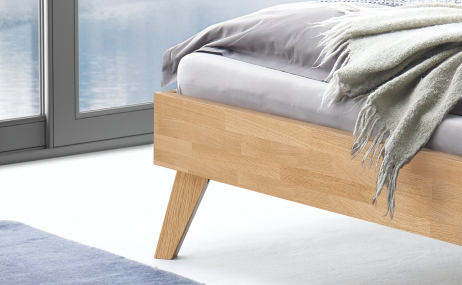 Detailfoto in zijaanzicht: modern bed van lichtgekleurd hout met schuine poten, grijs beddengoed en een beige deken aan het voeteneinde, voor een groot raam.