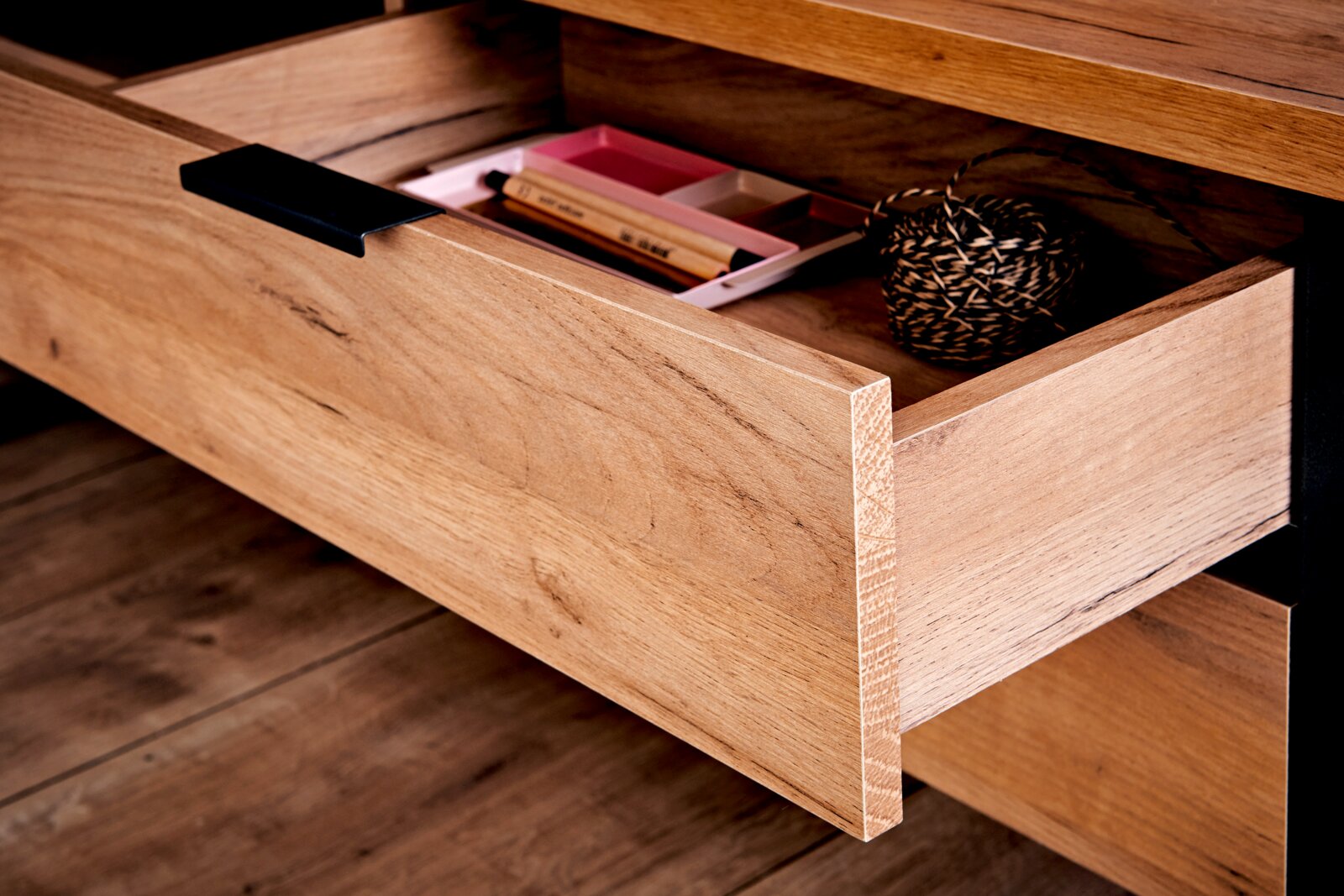 Nahaufnahme einer geöffneten Schublade eines Eck-Schreibtisches aus Holz mit schwarzem Griff, gefüllt mit Schreibutensilien und einer Schnurrolle.