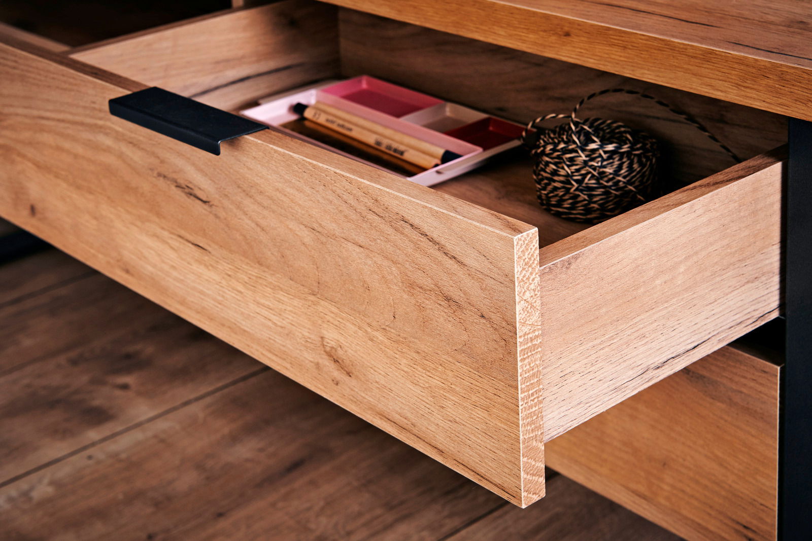 Nahaufnahme einer geöffneten Schublade eines Eck-Schreibtisches aus Holz mit schwarzem Griff, gefüllt mit Schreibutensilien und einer Schnurrolle.