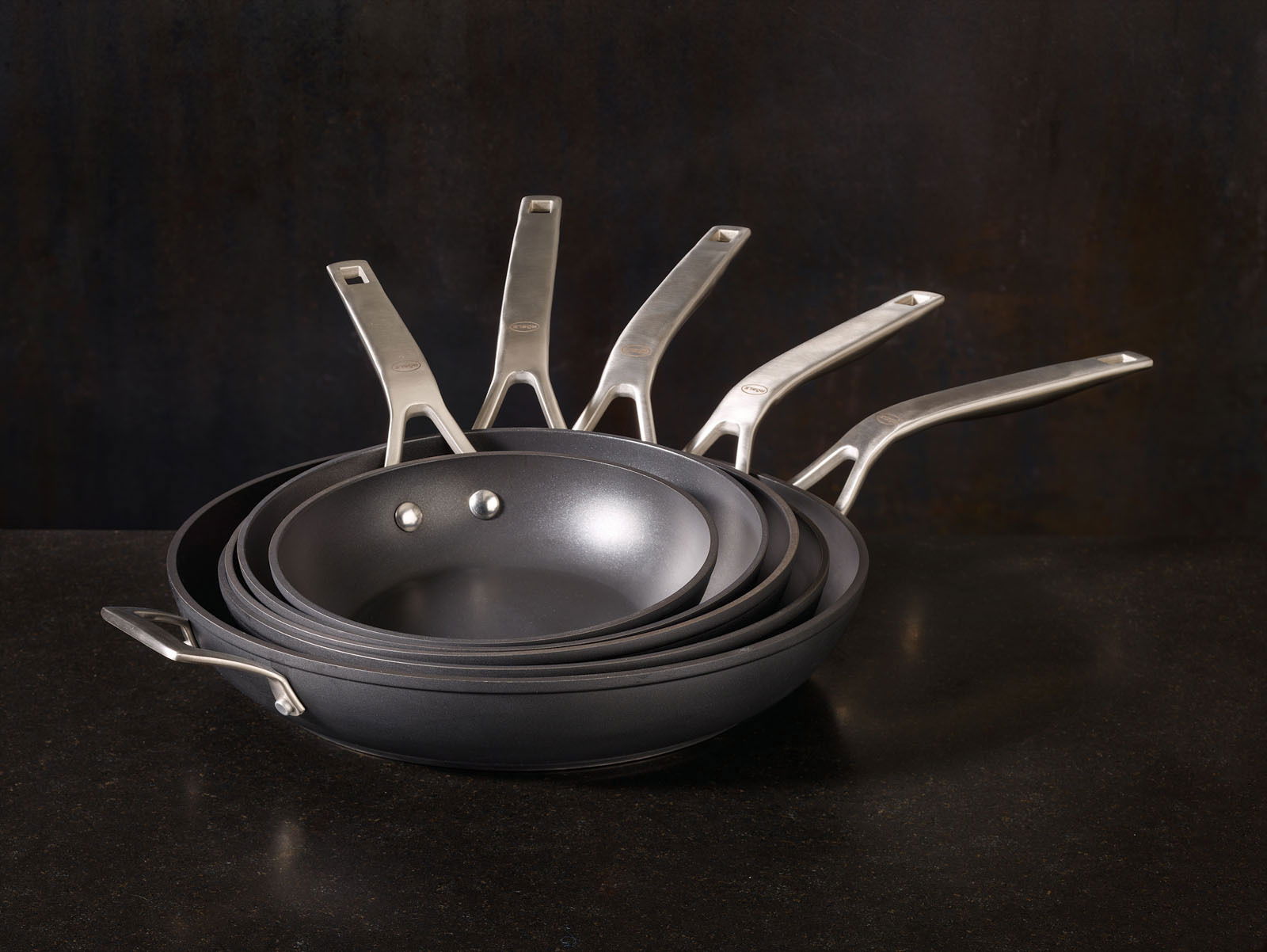 Ein Stapel von Bratpfannen der Serie Raise in verschiedenen Größen, von der Seite betrachtet, mit Edelstahlgriffen.