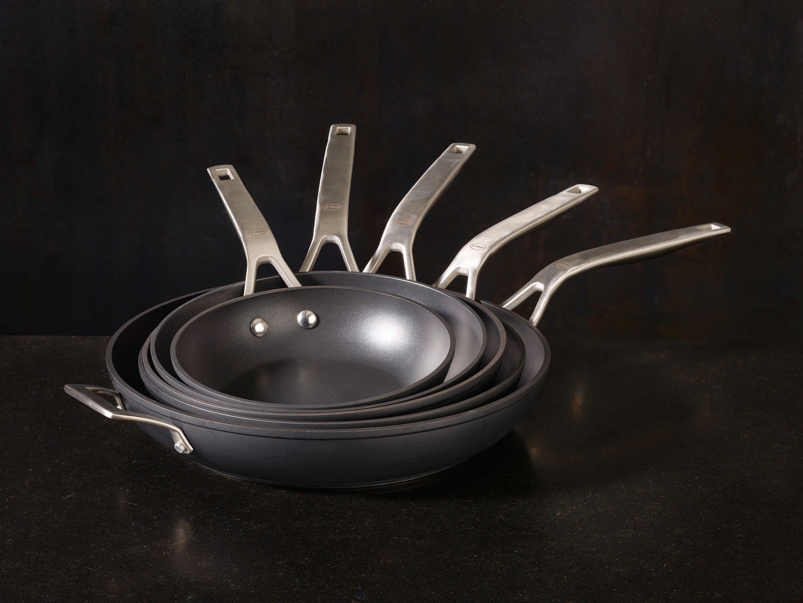 Ein Stapel von Bratpfannen der Serie Raise in verschiedenen Größen, von der Seite betrachtet, mit Edelstahlgriffen.