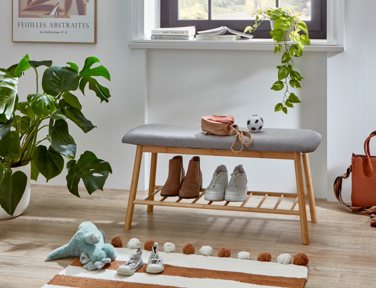 Gepolsterte Sitzbank mit grauem Stoffbezug und Holzgestell mit Schuhablage, frontal in einem hellen Flur unter einem Fenster inszeniert, mit Schuhen und kleinen Accessoires.