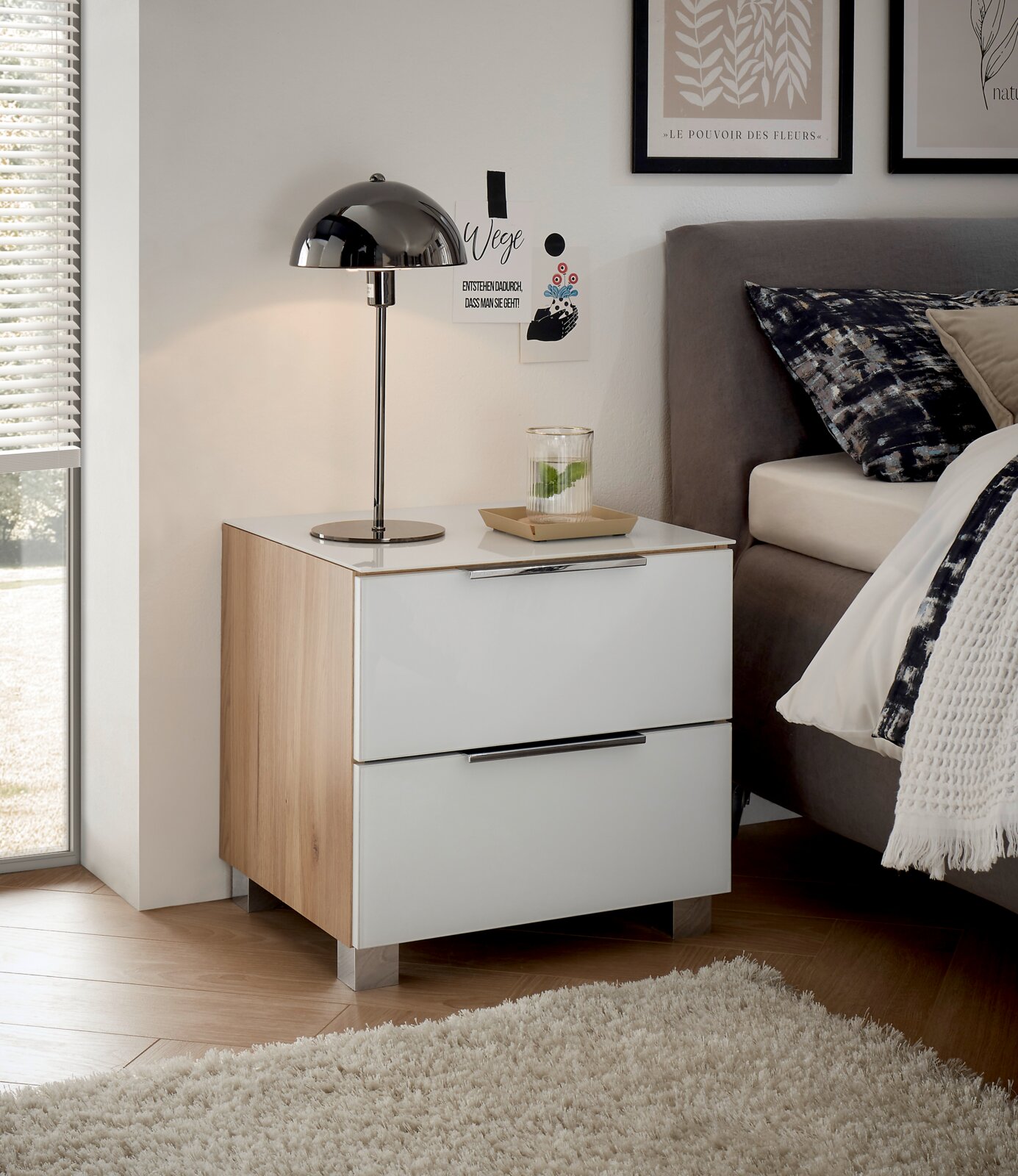 Moderne Nachtkommode mit zwei Schubladen in Holzoptik und weißem Hochglanz, seitliche Perspektive neben einem Bett mit Lampe und Dekoration.