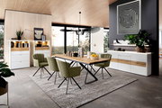 Moderner Essbereich mit einem Venjakob Esstisch 352PUH aus Holz und Metallgestell, umgeben von grünen Stühlen. Der Tisch steht auf einem Teppich in einem hellen Raum mit großen Fenstern und stilvoller Dekoration. Perspektive: seitliche Ansicht.