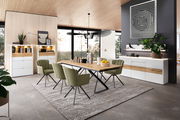 Moderner Essbereich mit einem Venjakob Esstisch 352PUH aus Holz und Metallgestell, umgeben von grünen Stühlen. Der Tisch steht auf einem Teppich in einem hellen Raum mit großen Fenstern und stilvoller Dekoration. Perspektive: seitliche Ansicht.