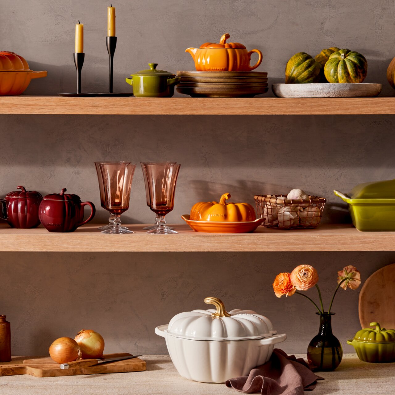 Weißer Kürbis-Bräter mit goldfarbenem Stiel, auf einem Tisch mit Zwiebeln und Blumen in einem Glasgefäß, aus seitlicher Perspektive.