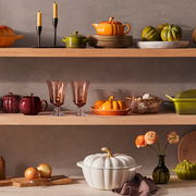 Weißer Kürbis-Bräter mit goldfarbenem Stiel, auf einem Tisch mit Zwiebeln und Blumen in einem Glasgefäß, aus seitlicher Perspektive.