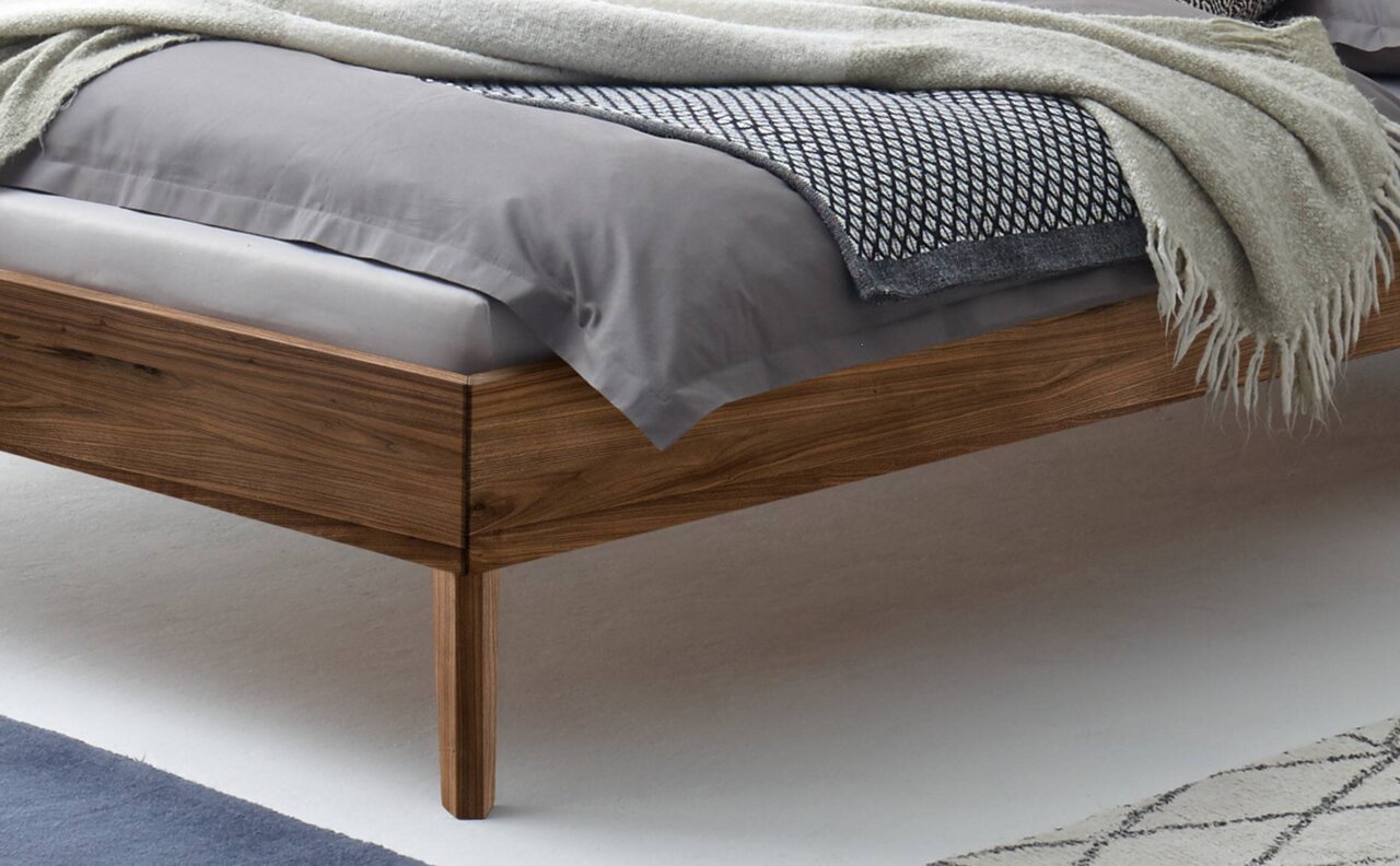 Close-up van een modern houten bedframe met grijs beddengoed en een deken, gezien vanaf de zijkant