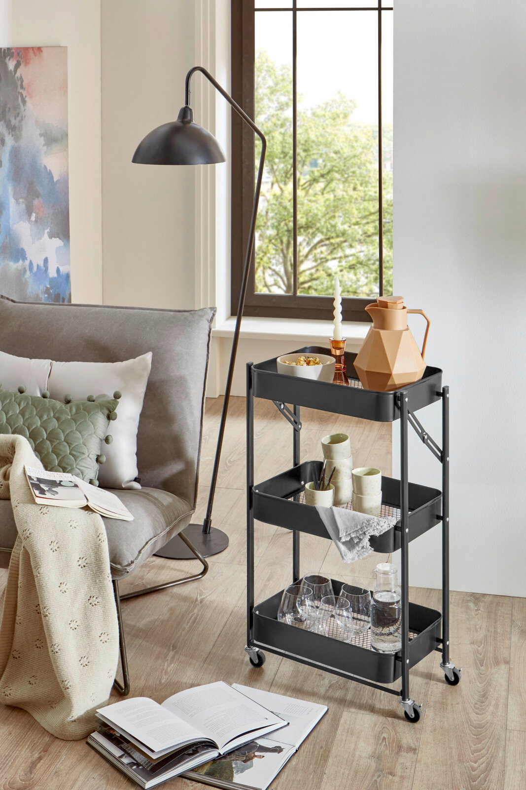 Schwarzer, dreistöckiger Servierwagen aus Metall mit Rollen und Gitterböden, seitliche Frontansicht im Wohnzimmer neben Sofa und Stehlampe, dekoriert mit Karaffe, Gläsern und Geschirr.