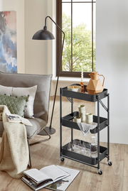 Schwarzer, dreistöckiger Servierwagen aus Metall mit Rollen und Gitterböden, seitliche Frontansicht im Wohnzimmer neben Sofa und Stehlampe, dekoriert mit Karaffe, Gläsern und Geschirr.