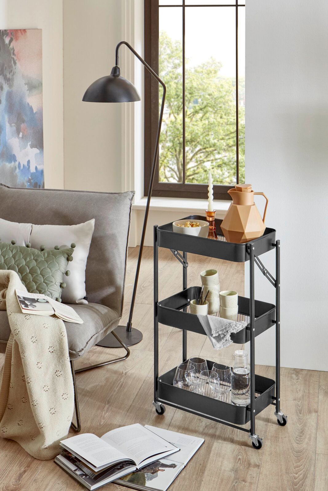 Schwarzer, dreistöckiger Servierwagen aus Metall mit Rollen und Gitterböden, seitliche Frontansicht im Wohnzimmer neben Sofa und Stehlampe, dekoriert mit Karaffe, Gläsern und Geschirr.