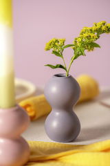 Fliederfarbene Keramikvase mit geschwungener Form, frontal fotografiert, mit gelben Blumen dekoriert.