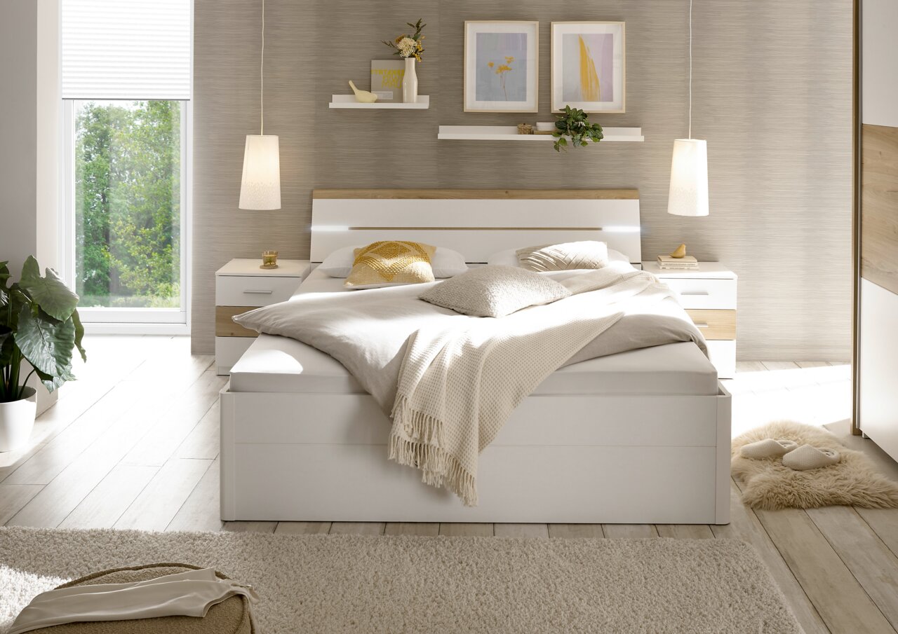 Modernes Schlafzimmer mit einem großen weißen Bett, flankiert von zwei Nachttischen. Das Bett ist mit neutralen Kissen und Decken dekoriert. Im Hintergrund sind zwei schwebende Regale mit Pflanzen und Bildern an der Wand. Die Perspektive zeigt das Schlafz