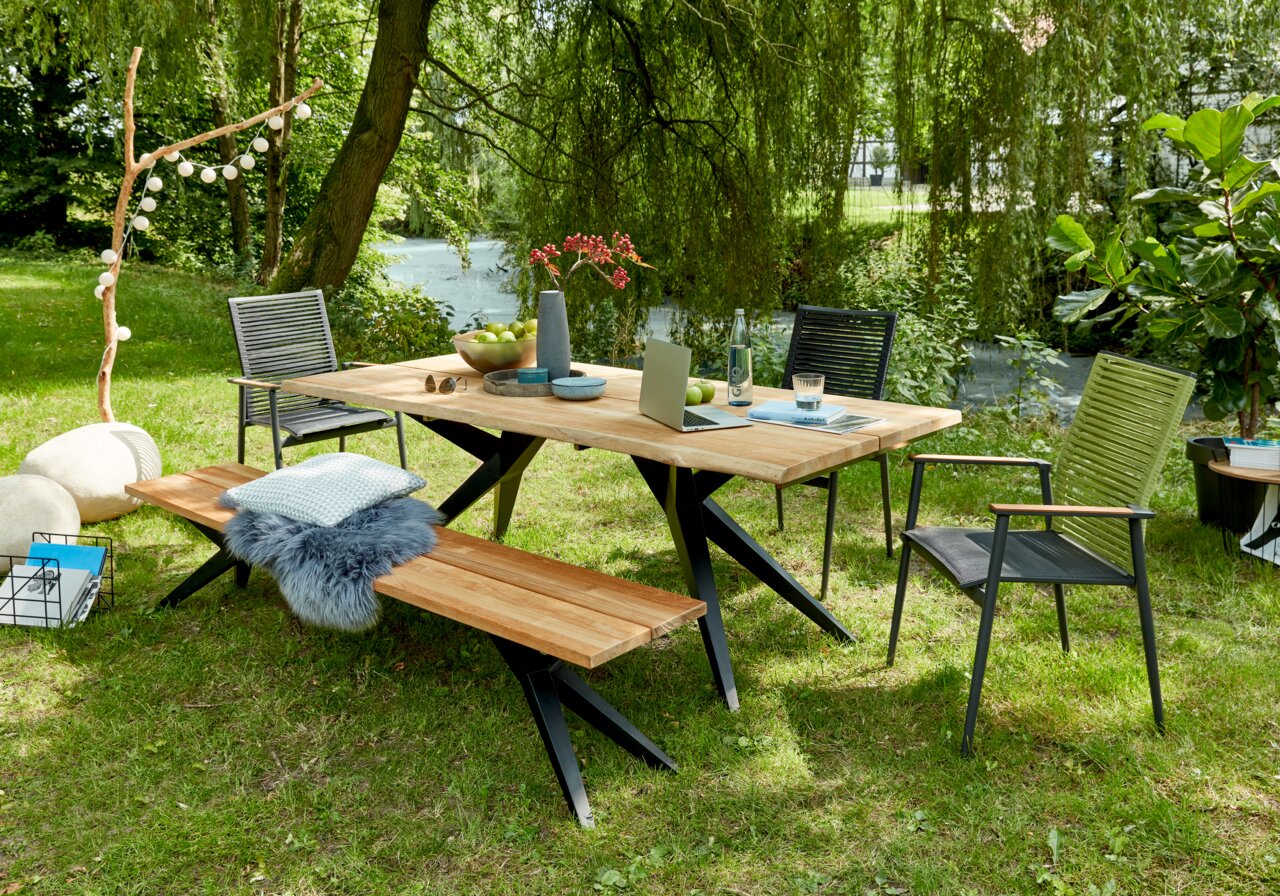 Holzbank im Garten neben einem Tisch, umgeben von Stühlen und grüner Natur, aus seitlicher Perspektive.