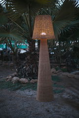 Outdoor-Stehleuchte aus Rattan in einem Garten bei Dämmerung, frontal fotografiert.