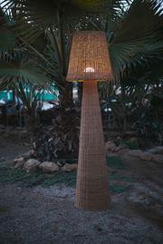 Outdoor-Stehleuchte aus Rattan in einem Garten bei Dämmerung, frontal fotografiert.
