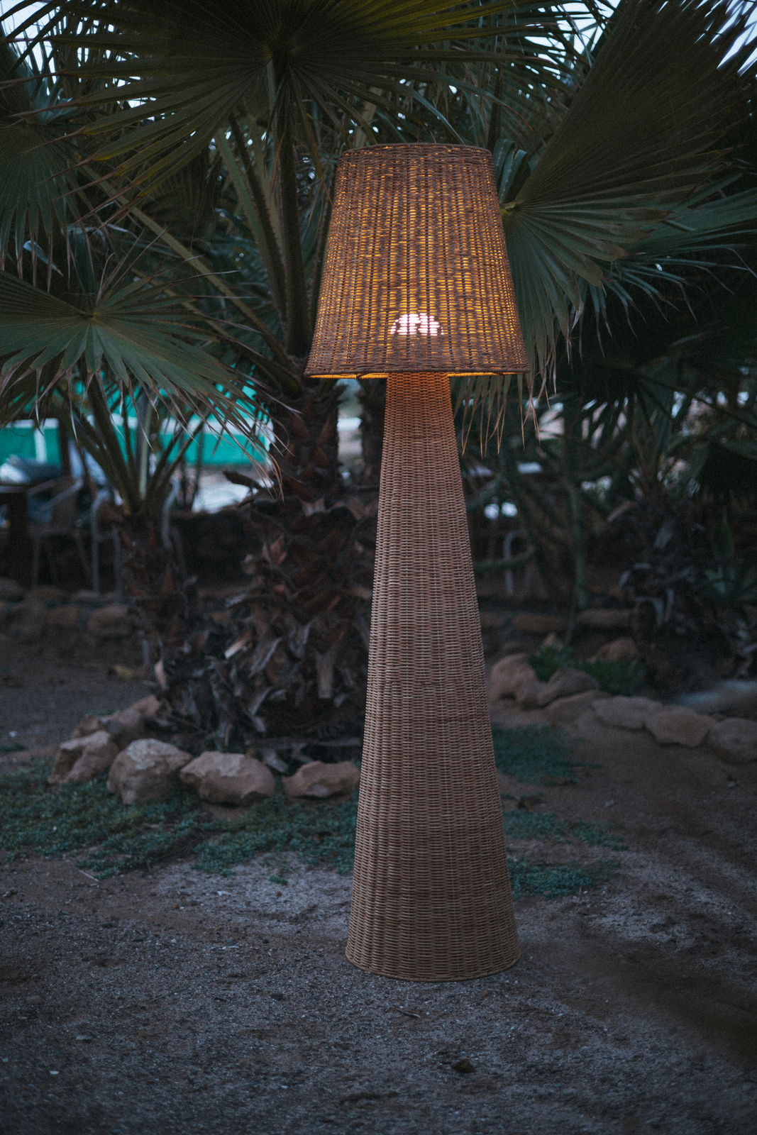 Outdoor-Stehleuchte aus Rattan in einem Garten bei Dämmerung, frontal fotografiert.