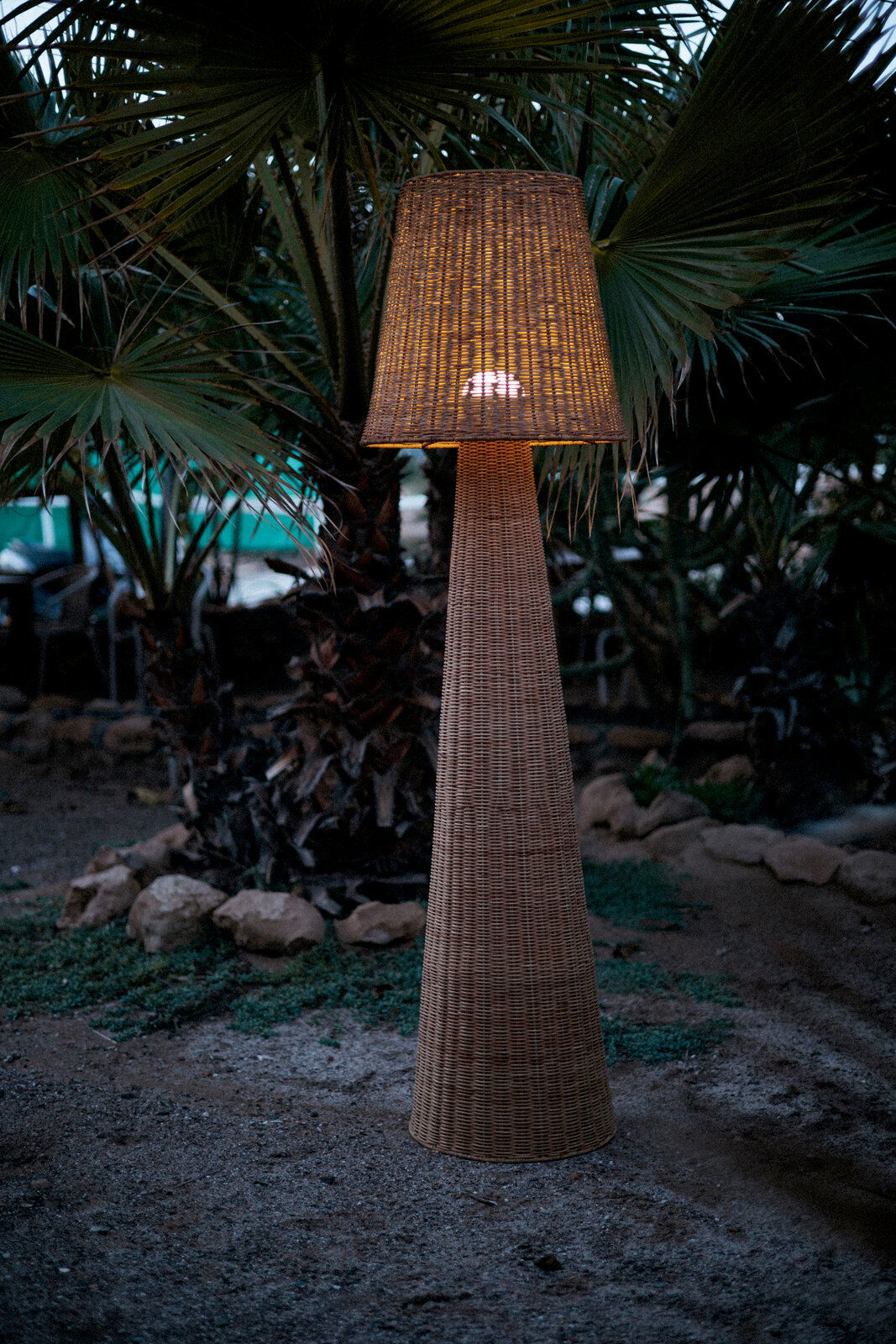 Outdoor-Stehleuchte aus Rattan in einem Garten bei Dämmerung, frontal fotografiert.