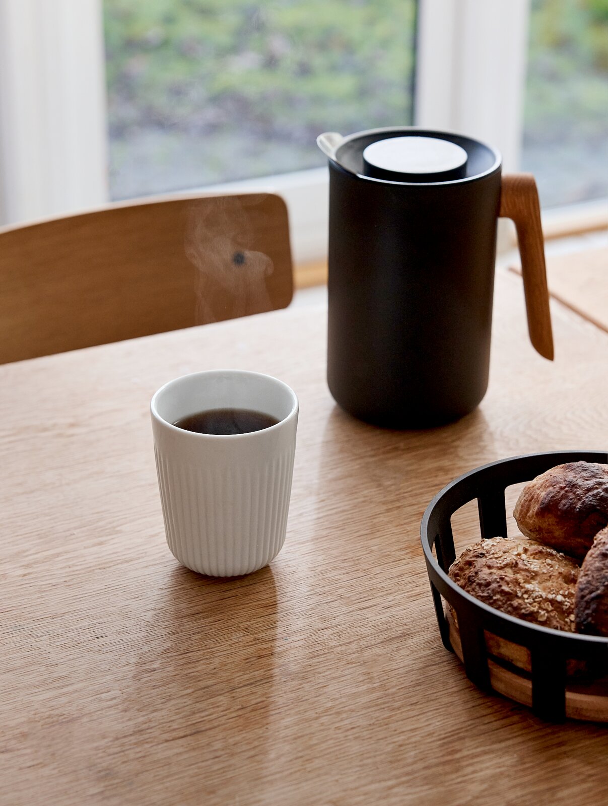 Zwarte thermoskan met houten handgreep op een houten tafel naast een witte koffiekop en een broodmand, gefotografeerd schuin van voren op tafelhoogte bij het raam.
