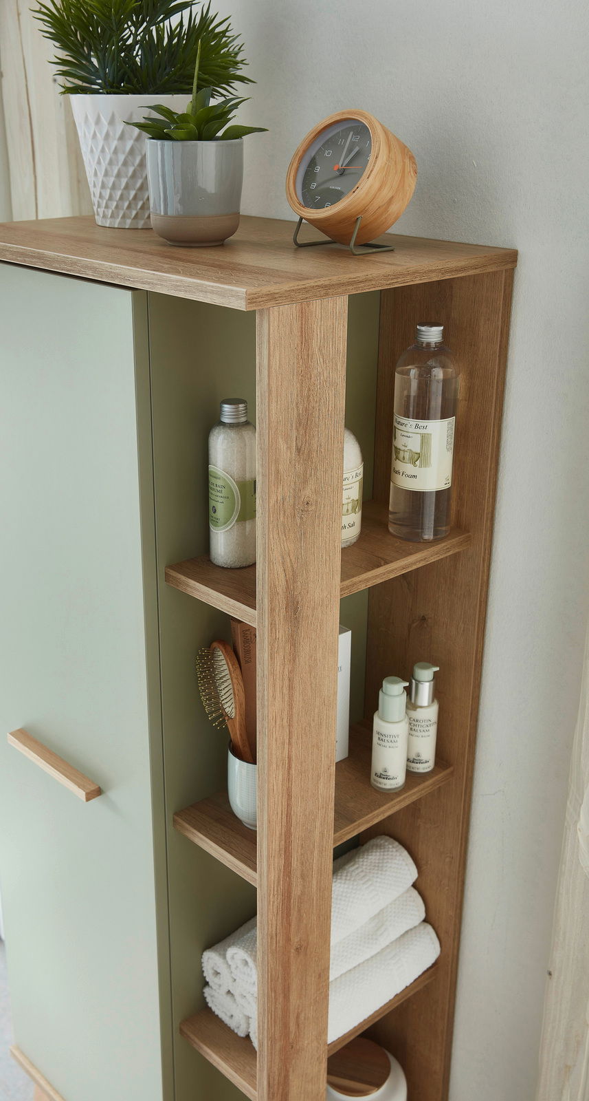 Midischrank mit Holzoptik und grüner Tür, seitlich fotografiert, mit dekorativen Pflanzen, Uhr und Pflegeprodukten auf den Regalen.