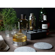 Eiswürfelform mit eckigen Fächern, gefüllt mit klaren Eiswürfeln, im Vordergrund ein Glas mit Getränk und Eiswürfeln, im Hintergrund Flaschen und ein Cocktailshaker, seitliche Perspektive