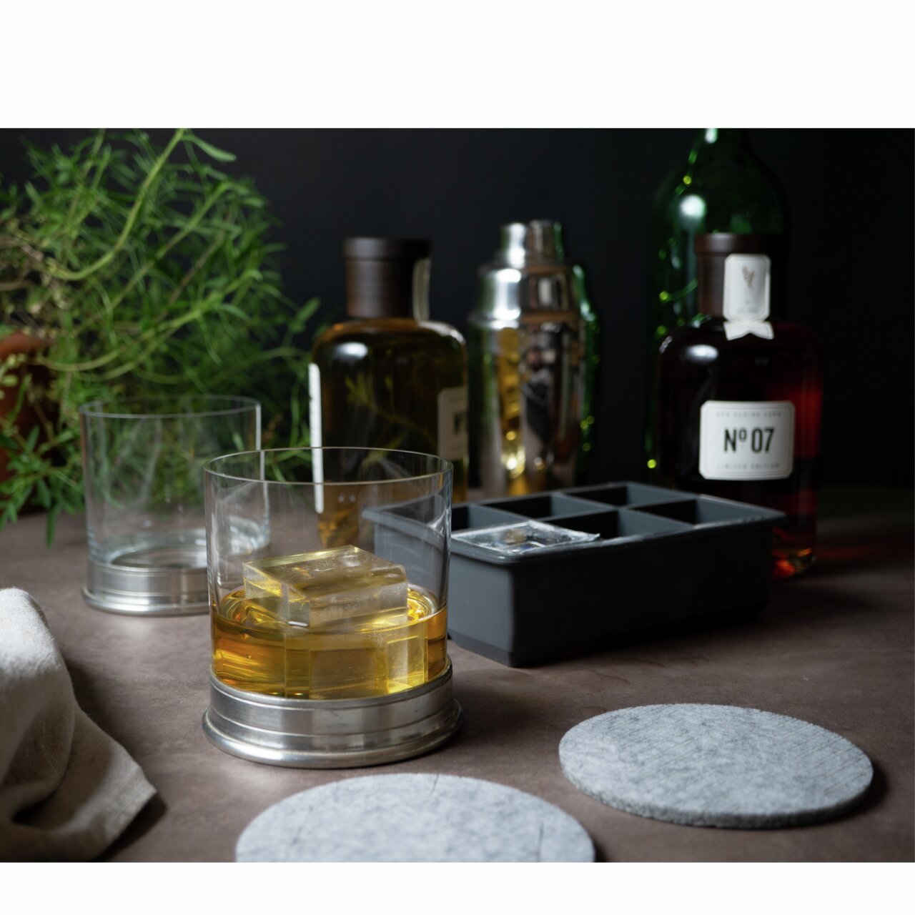 Eiswürfelform mit eckigen Fächern, gefüllt mit klaren Eiswürfeln, im Vordergrund ein Glas mit Getränk und Eiswürfeln, im Hintergrund Flaschen und ein Cocktailshaker, seitliche Perspektive