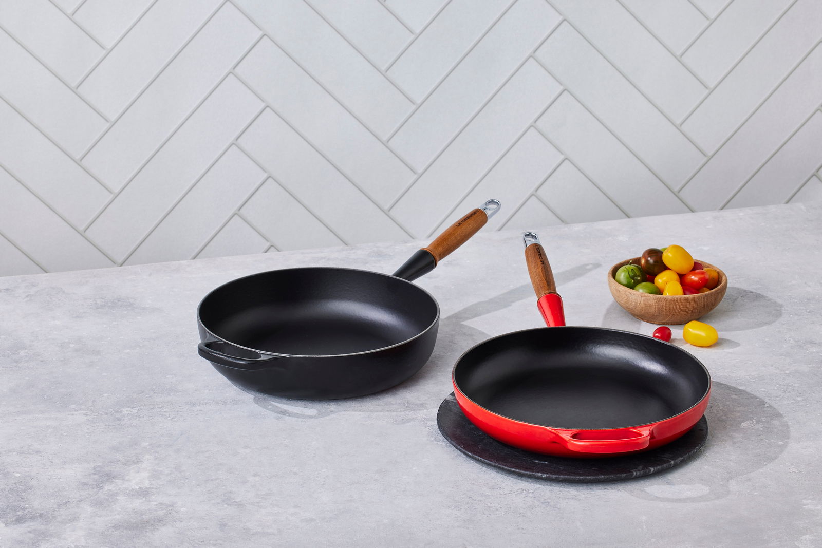 Zwei Bratpfannen auf einer Küchenarbeitsplatte, eine in Kirschrot und eine in Schwarz, mit Holzgriffen, aus der Vogelperspektive fotografiert.