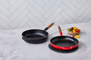 Zwei Bratpfannen auf einer Küchenarbeitsplatte, eine in Kirschrot und eine in Schwarz, mit Holzgriffen, aus der Vogelperspektive fotografiert.