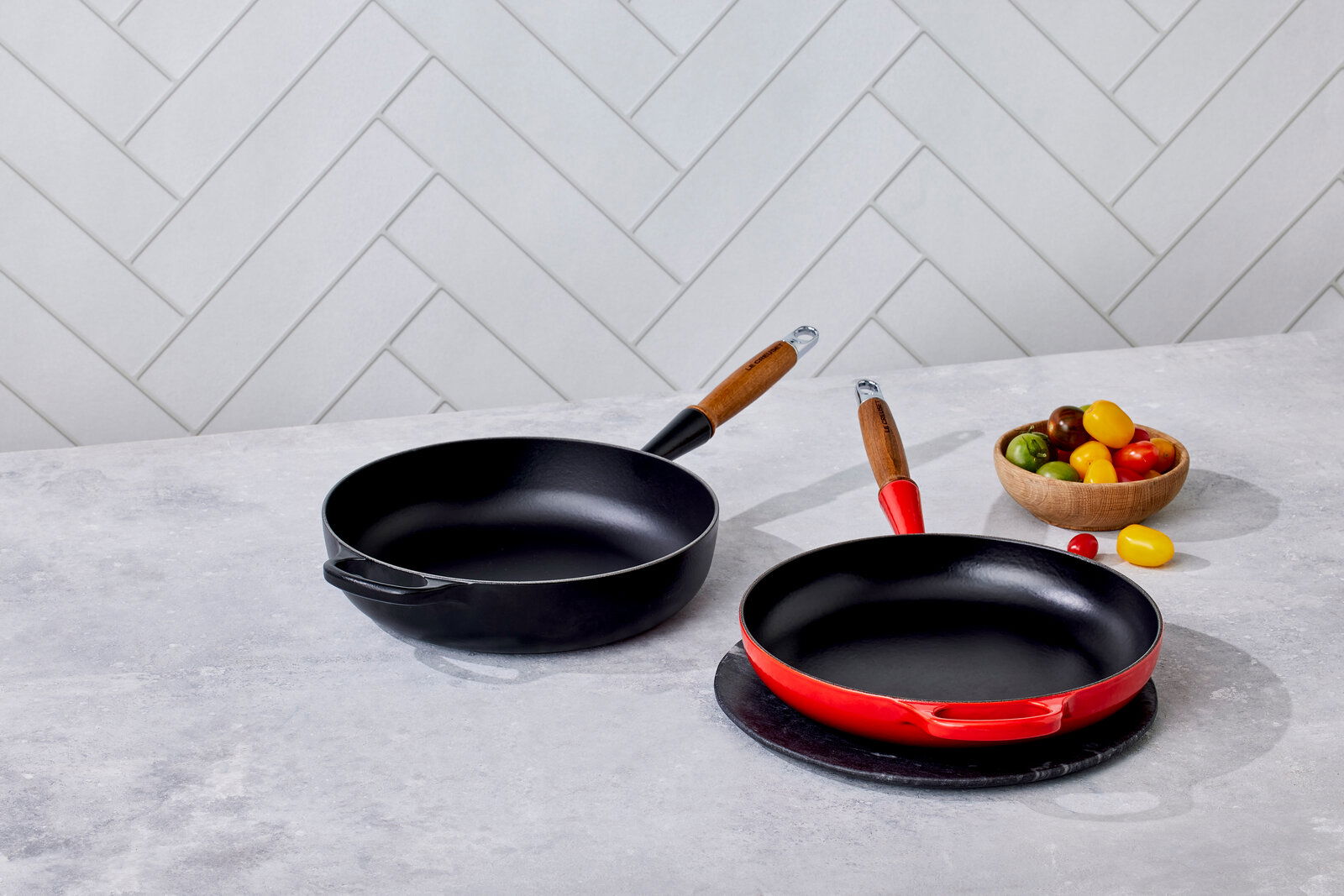 Zwei Bratpfannen auf einer Küchenarbeitsplatte, eine in Kirschrot und eine in Schwarz, mit Holzgriffen, aus der Vogelperspektive fotografiert.