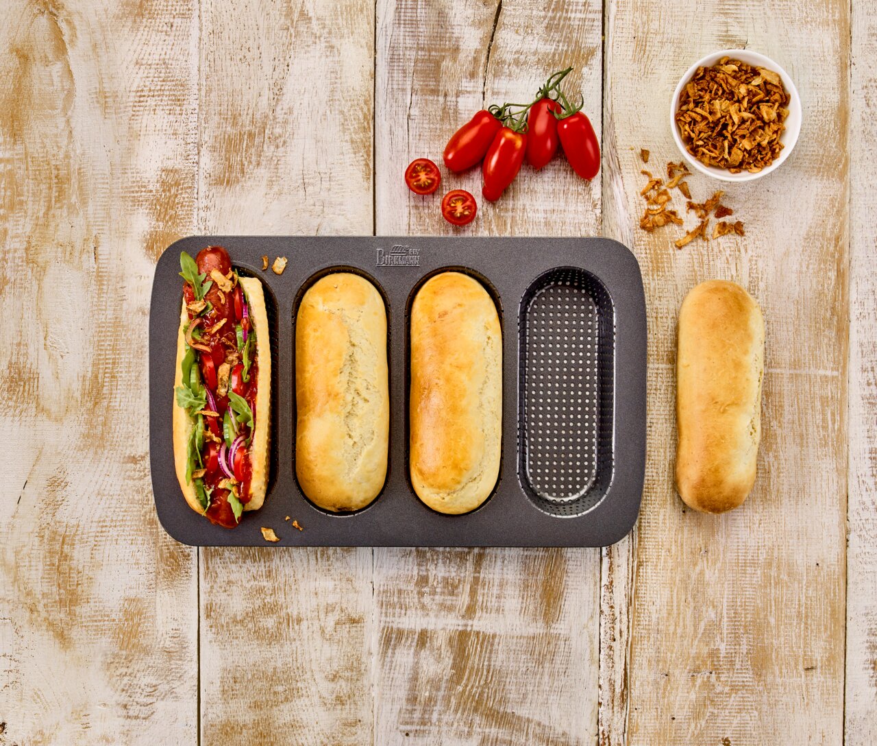 Vogelperspektive auf ein Hot Dog Brötchen-Blech mit drei gebackenen Brötchen und einem belegten Hot Dog, umgeben von frischen Tomaten und Röstzwiebeln auf einem Holztisch.