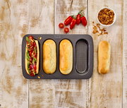 Vogelperspektive auf ein Hot Dog Brötchen-Blech mit drei gebackenen Brötchen und einem belegten Hot Dog, umgeben von frischen Tomaten und Röstzwiebeln auf einem Holztisch.