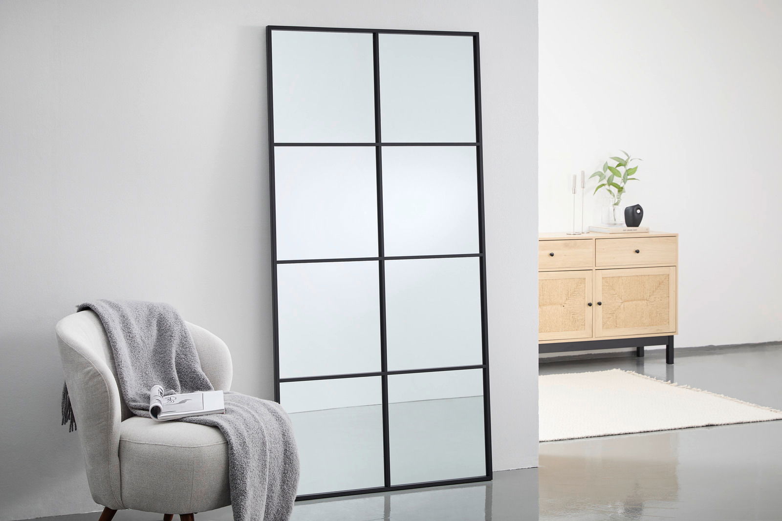 Großer rechteckiger Spiegel mit schwarzem Rahmen, in neun Abschnitte unterteilt, lehnt an einer weißen Wand. Perspektive von schräg vorne.