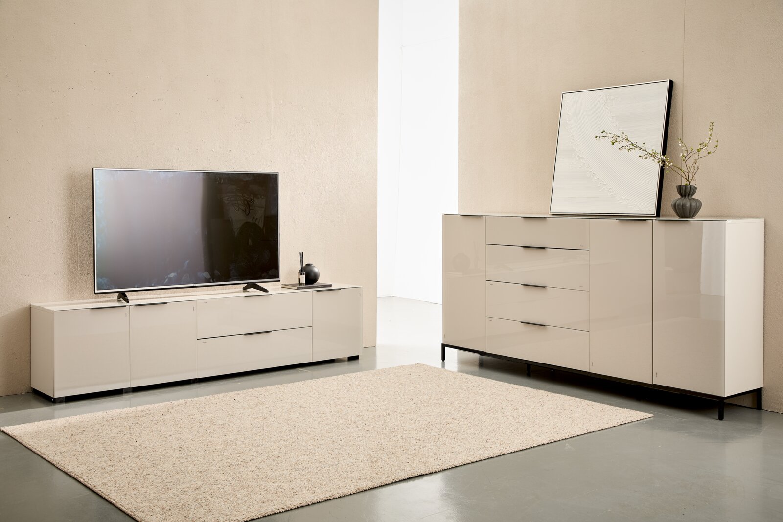 Wohnzimmer-Set mit modernem TV-Lowboard und Sideboard in Beige/Creme mit schwarzen Griffen und Metallfüßen, frontal in leicht schräger Perspektive fotografiert; links ein Fernseher auf dem Lowboard, rechts Deko auf dem Sideboard