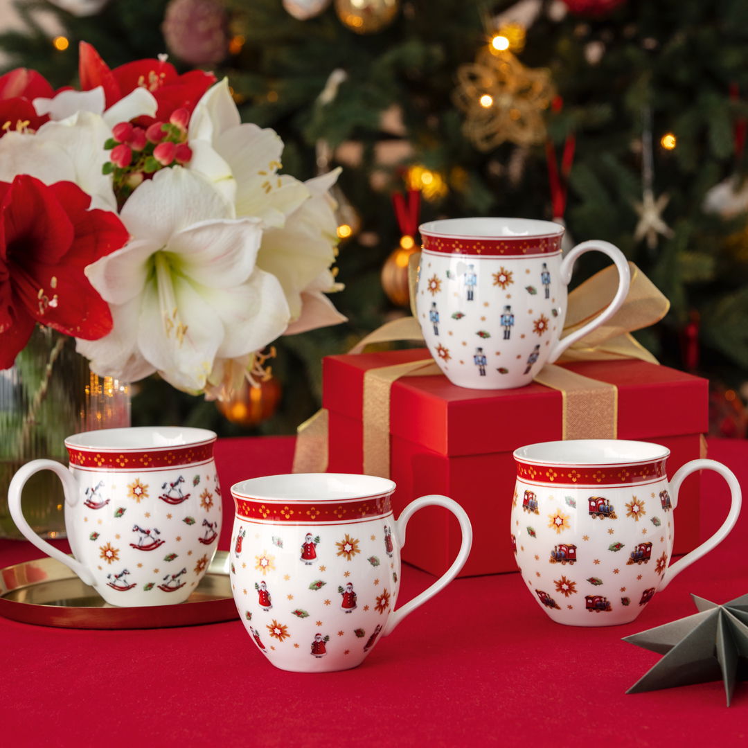 Vier festliche Jubiläums-Becher mit weihnachtlichen Motiven, präsentiert auf einem Tisch mit rotem Tischtuch, im Hintergrund ein geschmückter Weihnachtsbaum. Perspektive: Frontalansicht.