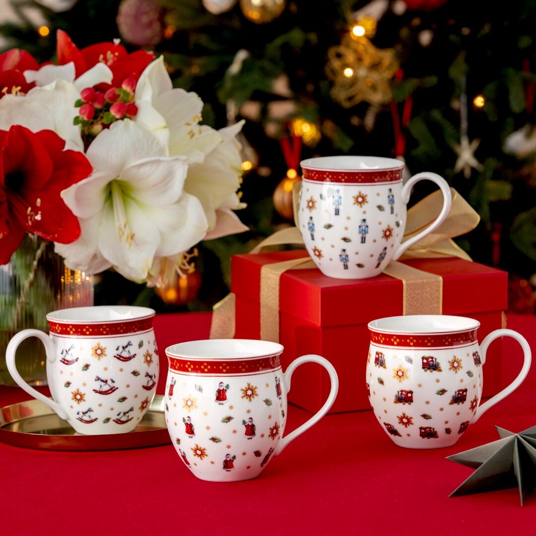 Villeroy & Boch Jubiläums-Becher-Set 4tlg Vier festliche Jubiläums-Becher mit weihnachtlichen Motiven, präsentiert auf einem Tisch mit rotem Tischtuch, im Hintergrund ein geschmückter Weihnachtsbaum. Perspektive: Frontalansicht.