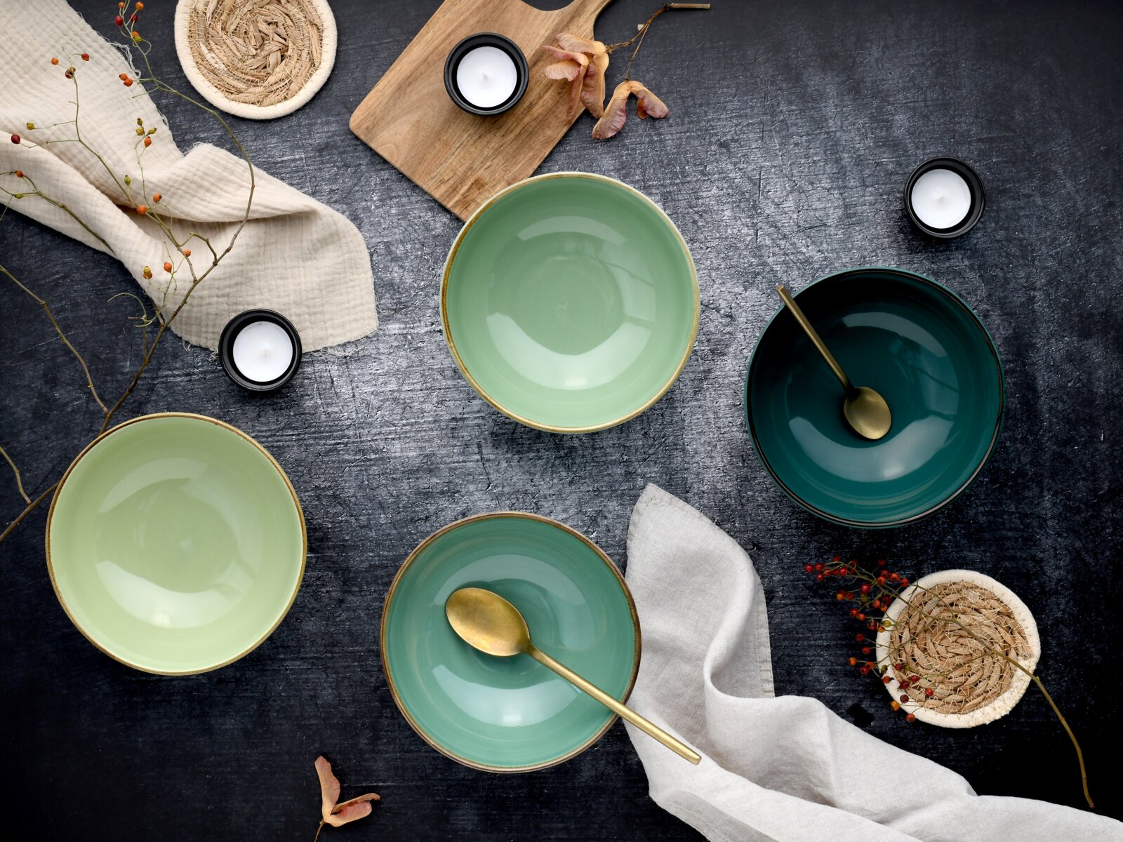 Drei glasierte Keramikschalen in Mint- und Petrolgrün mit goldfarbenen Löffeln, dekoriert mit Leinentuch, Teelichtern und Naturdetails auf dunkler Oberfläche, fotografiert in Draufsicht (Flatlay)