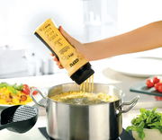 Hand hält eine Flasche Crepes-/Pfannkuchen-Mix über einem Topf, aus der der Teig in kochendes Wasser fließt. Perspektive von der Seite.