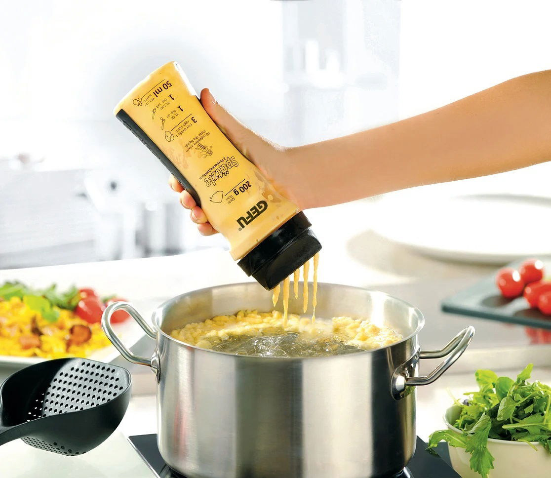 Hand hält eine Flasche Crepes-/Pfannkuchen-Mix über einem Topf, aus der der Teig in kochendes Wasser fließt. Perspektive von der Seite.