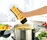 Hand hält eine Flasche Crepes-/Pfannkuchen-Mix über einem Topf, aus der der Teig in kochendes Wasser fließt. Perspektive von der Seite.