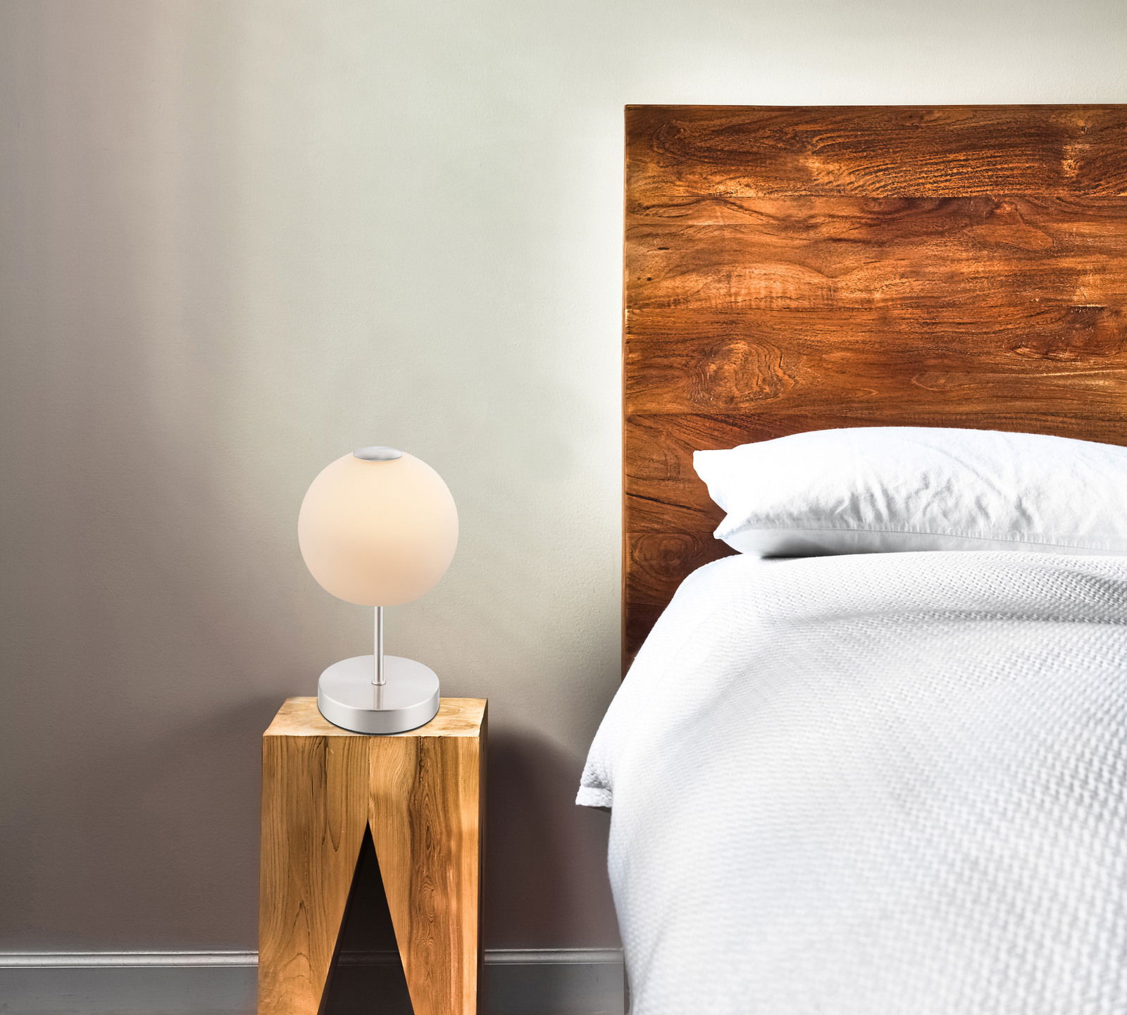 Moderne Tischleuchte aus mattem Nickel auf einem Holztisch neben einem Bett, seitliche Perspektive.