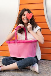 Frau sitzt lächelnd mit einem pinken Carrybag auf dem Schoß und einem herzförmigen Lutscher in der Hand vor einer orangefarbenen Wand. Frontalansicht.