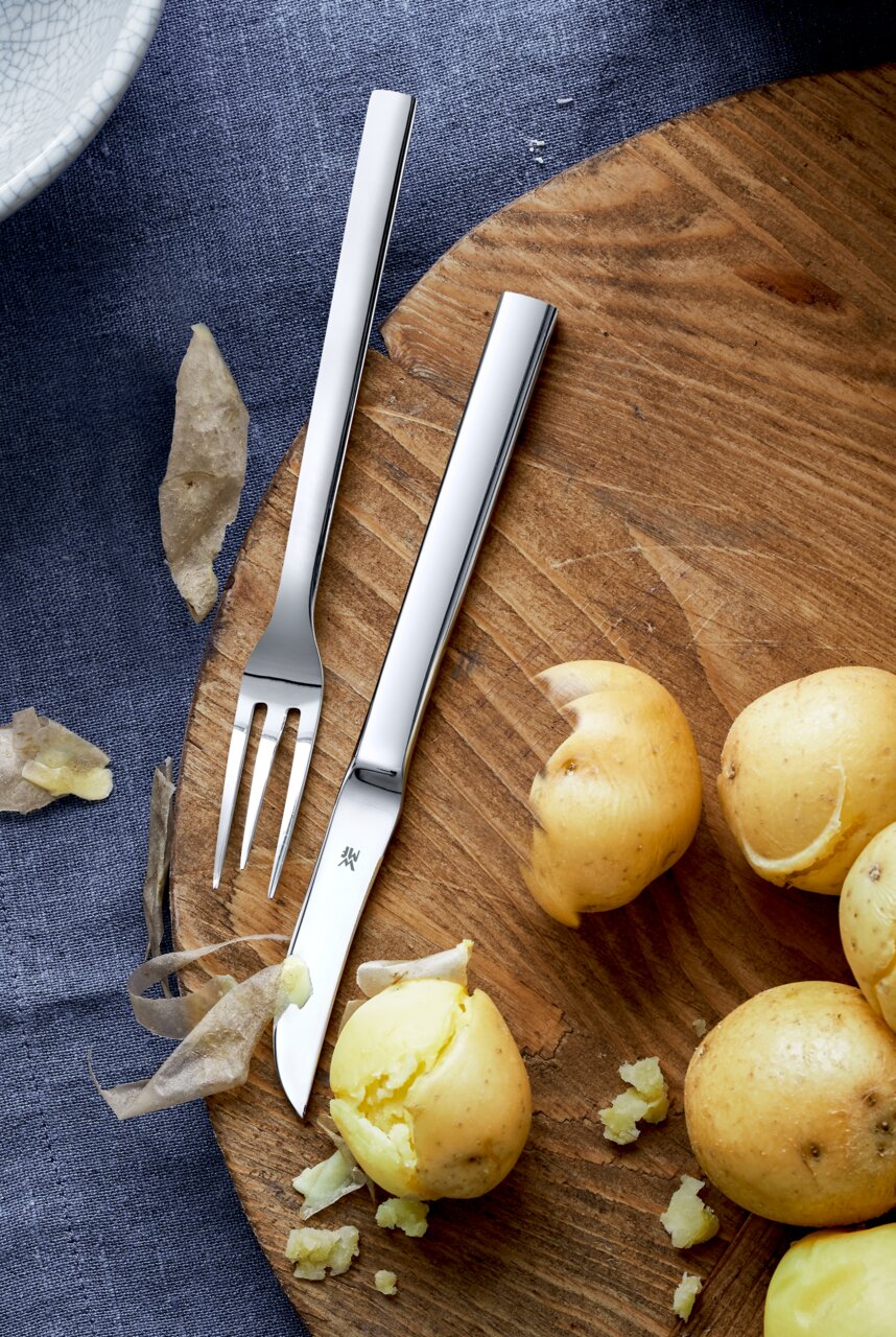 Vogelperspektive auf ein Pellkartoffel-Set mit Messer und Gabel auf einem Holzbrett, umgeben von geschälten Kartoffeln und Schalen.