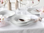Elegantes weißes Porzellan-Tafelservice mit tiefem Teller und flachem Teller, dekorativ gedeckter Tisch mit Gläsern, Besteck und Blumen, perspektivisch in leichter Schrägansicht von vorne