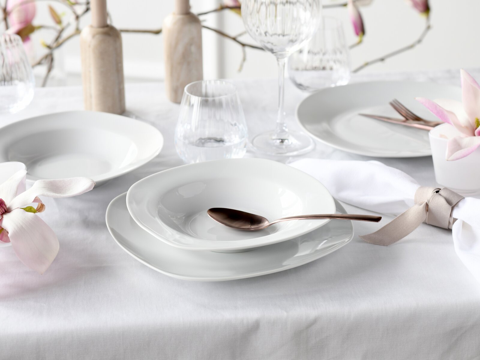 Elegantes weißes Porzellan-Tafelservice mit tiefem Teller und flachem Teller, dekorativ gedeckter Tisch mit Gläsern, Besteck und Blumen, perspektivisch in leichter Schrägansicht von vorne