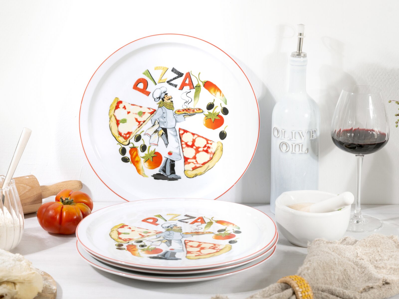 Pizza-Teller-Set aus weißem Porzellan mit rotem Rand und farbigem Pizza-Motiv (Schriftzug „PIZZA“, Pizzastücke, Koch, Tomaten und Oliven), in schräger Frontansicht auf gedecktem Küchentisch; ein Teller steht im Hintergrund, mehrere liegen gestapelt im Vor
