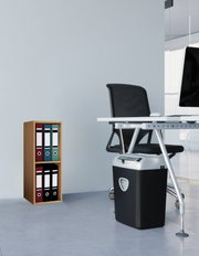 Schwarzer Aktenvernichter unter einem modernen Büroschreibtisch, rechts im Bild aus leicht seitlicher Perspektive, mit Ordnerregal im Hintergrund