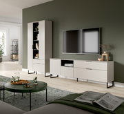 Modernes Wohnzimmer-Setup mit hellbeigem TV-Lowboard und passendem Hochschrank auf schwarzen Metallfüßen, wandmontiertem Fernseher und Deko vor olivgrüner Wand, perspektivisch in leichter Schrägansicht von vorn.