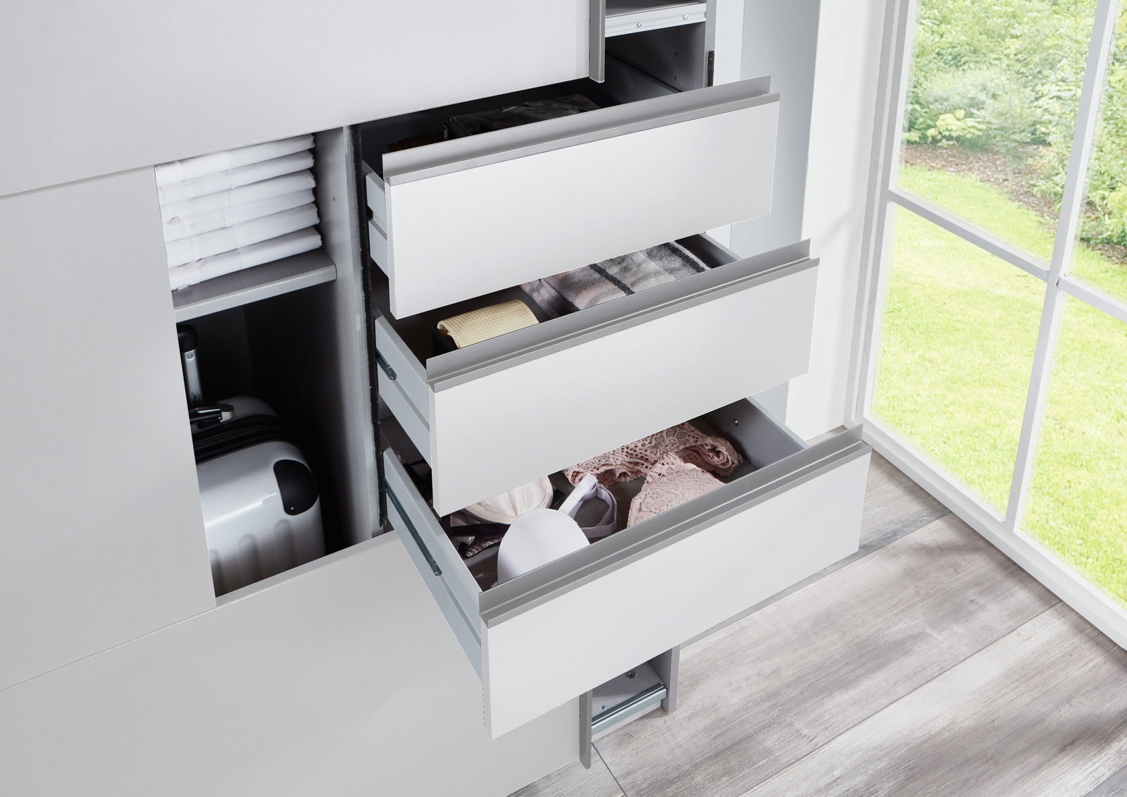 Nahaufnahme eines Schwebetürenschranks mit geöffneten Schubladen, die Kleidung und Accessoires zeigen, neben einem Fenster mit Blick ins Grüne.